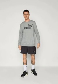 Harmaa huppari, jossa on musta "PUMA" -logo, yhdistettynä mustiin shortseihin. Mallilla on valkoiset sukat ja mustat urheilukengät vihreillä yksityiskohdilla.