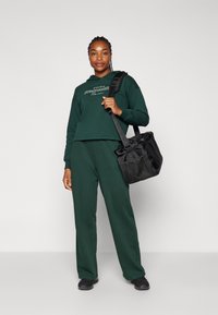Felpa verde a vita corta con testo bianco, abbinata a pantaloni verdi coordinati. Borsa a rete nera appesa alla spalla. Scarpe nere completano il look.