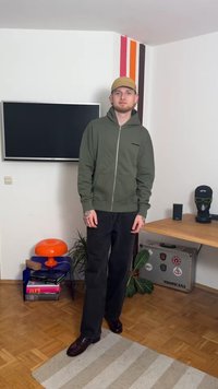 Olive green zip-up hoodie with front pockets, black pants, and brown shoes, standing on a wooden floor with a striped wall in the background.