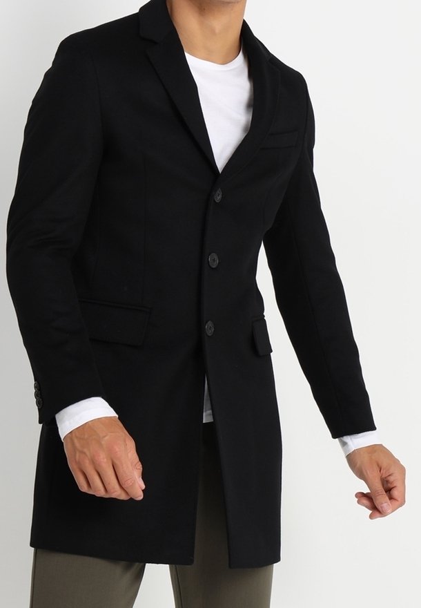 Un uomo in un cappotto nero aderente gesticola con la mano destra, indossando una camicia bianca e pantaloni verde oliva, su uno sfondo semplice.