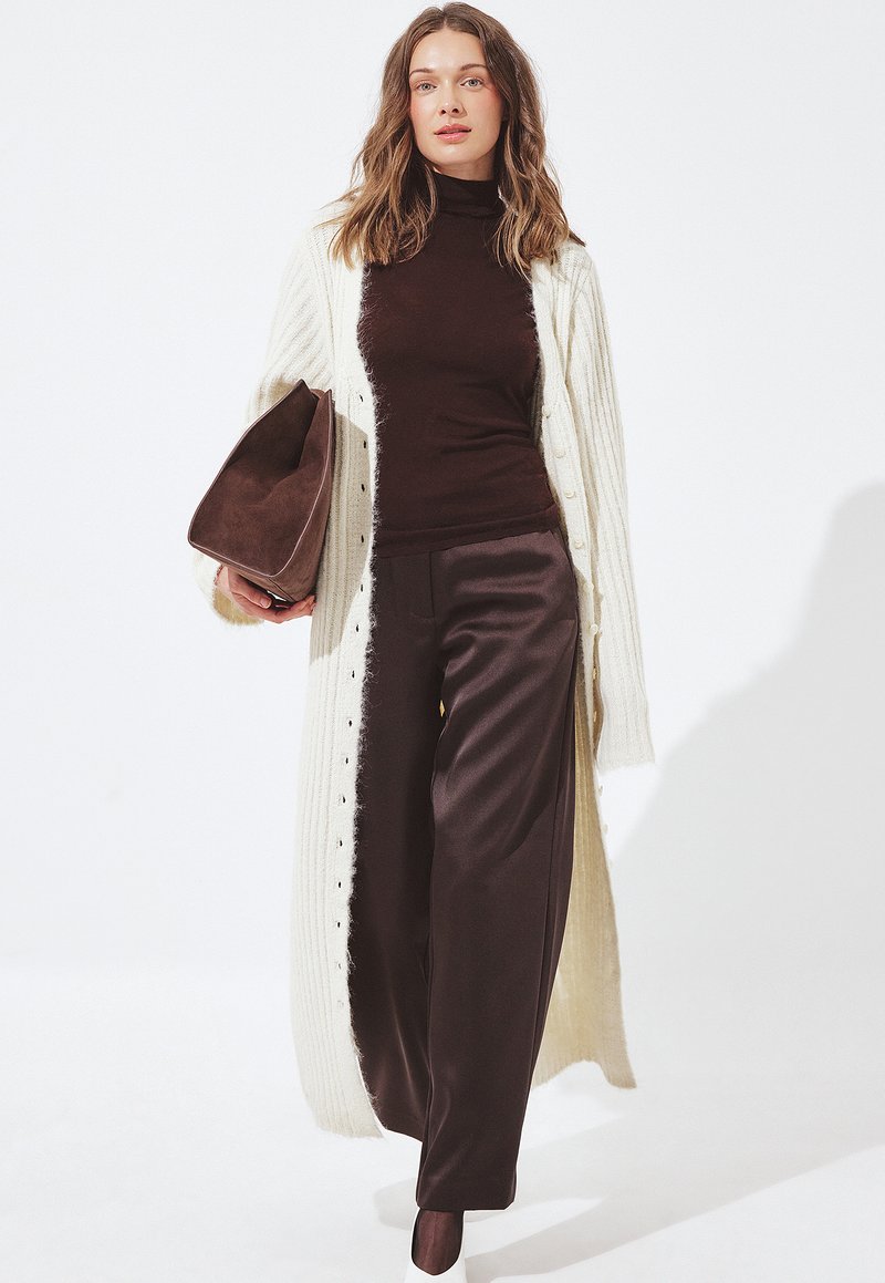 Donna che indossa un lungo cardigan color crema, un dolcevita marrone scuro, pantaloni a gamba larga abbinati, scarpe bianche e tiene una grande borsa in suede marrone.