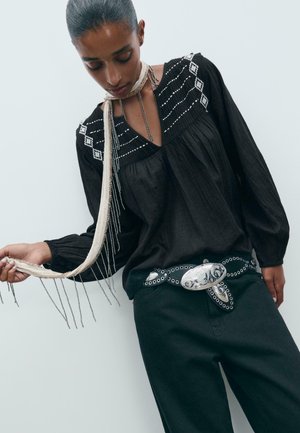 Blusa nera in tessuto testurizzato, con cuciture decorative bianche e scollatura a V. Abbinata a pantaloni scuri e una cintura con dettagli argentati.