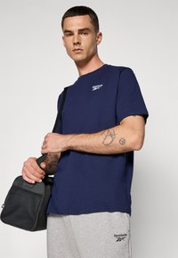 Mørkeblå bomulds t-shirt med rund hals og et lille hvidt Reebok logo; grå træningsbukser med logo på benet; sort skuldertaske.