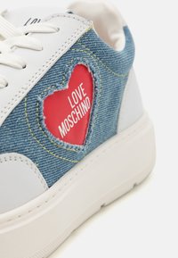 Jeansneakers med en vit sula, ett rött hjärtutsnitt med texten "LOVE MOSCHINO" och gula sömnadsdetaljer på blått tyg.