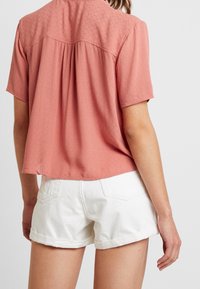 Blouse à manches courtes couleur corail clair avec un design texturé côtelé et un dos plissé, associée à un short en jean blanc retroussé.