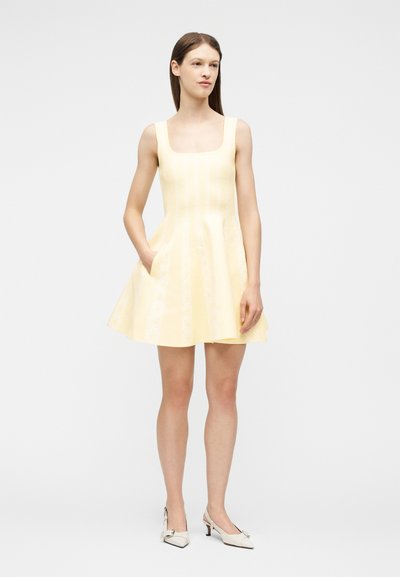 Γυναίκα όρθια με αμάνικο παλ χρωματιστό κίτρινο φόρεμα με τετράγωνο ντεκολτέ και λευκά παπούτσια με χαμηλό τακούνι.