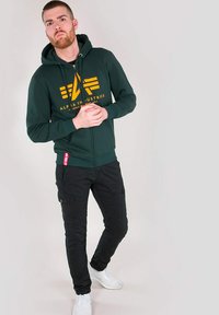 Felpa verde con zip e logo giallo, abbinata a pantaloni cargo neri e sneakers bianche. La felpa presenta una tasca a marsupio e lacci.