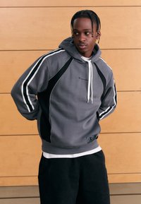 Grå huvtröja med svarta detaljer och vita ränder, utrustad med en känguruficka och elastiska muddar. Broderad logotyp på bröstet.