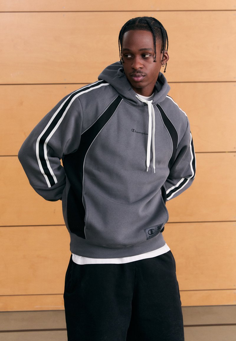 Grå huvtröja med svarta detaljer och vita ränder, utrustad med en känguruficka och elastiska muddar. Broderad logotyp på bröstet.