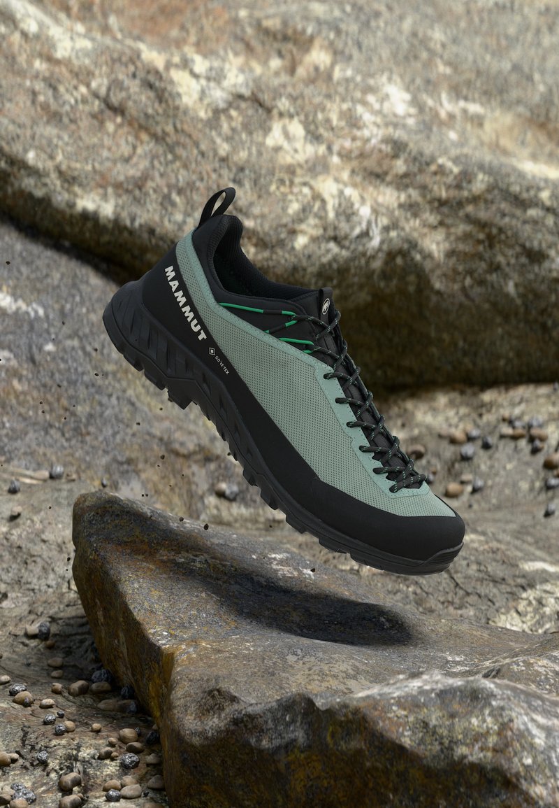 Πράσινο και μαύρο παπούτσι πεζοπορίας Mammut με Gore-Tex, εμφανιζόμενο πάνω από πετρώδες εξωτερικό έδαφος, τονίζοντας την ανθεκτική σόλα και τις λεπτομέρειες των κορδονιών.