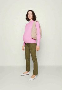 Różowy prążkowany sweter, oliwkowe spodnie cargo i białe baleriny. Modelka nosi ponad jedno ramię teksturowaną beżową torbę.