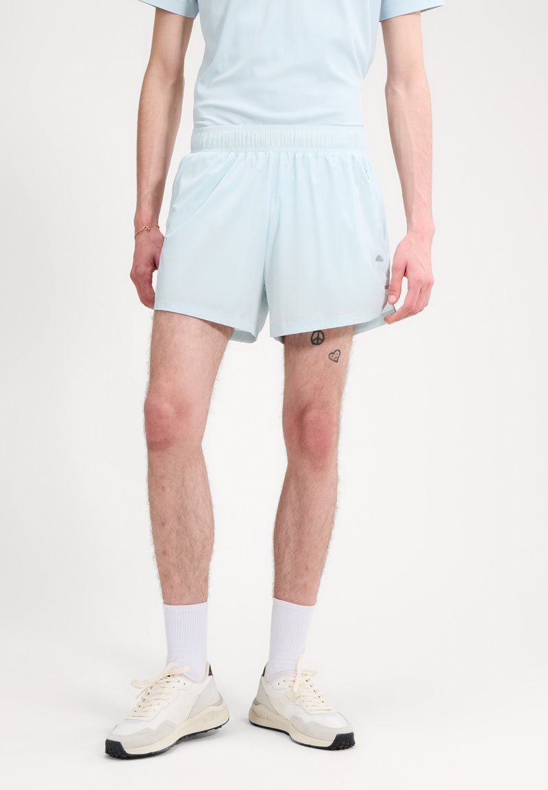 Άνδρας που φοράει ανοιχτό μπλε σορτς, λευκές κάλτσες και λευκά αθλητικά παπούτσια, δείχνοντας τη χειρονομία της ειρήνης και τα τατουάζ με καρδιά στο δεξί μηρό.