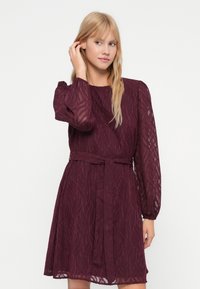 Blonde sieviete, kas pielāgo mati, ģērbusies garajā, līdz ceļiem, caurspīdīgajā bordo kleitā ar teksturētu lapu rakstu un jostu vidukļa apvidū.