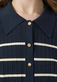 Marineblå stickad skjorta med vita horisontella ränder, med klassisk krage och dekorerade knappar med pärl-liknande mittdelar och rhinestones. 