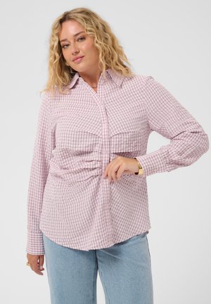Femme blonde portant une chemise à carreaux rose clair boutonnée et un jean bleu clair, posant avec une main près de la taille.