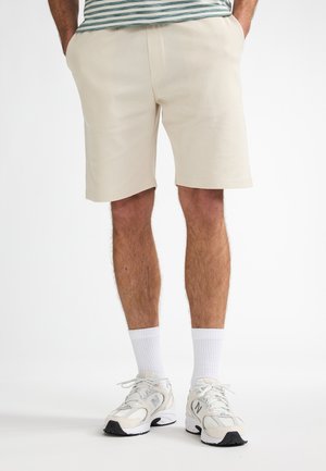 Beige, strukturierte Shorts mit lässiger Passform und Seitentaschen. Kombiniert mit weißen Socken und hellen Sneakern.