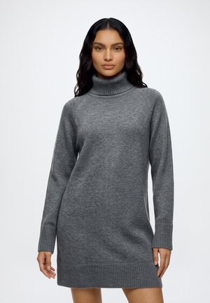 Un mannequin aux longs cheveux foncés porte une robe-pull grise côtelée à col roulé, sur un fond clair et uni.