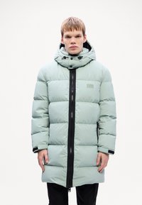 Padded parka u svijetlozelenoj boji sa kapuljačom na čvrstu uže, crnim prednjim zipom i elastičnim manžetama. Džepovi s oblogom od flis-a sa strana.