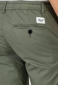 Bermudas verde oliva hechas de un tejido suave, con un bolsillo trasero que tiene un botón negro y una etiqueta blanca con el nombre de la marca cosida arriba.