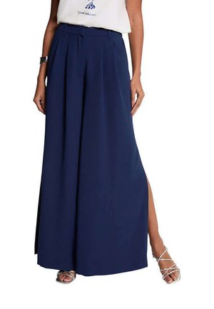 Kvinde iført marineblå, vidde bukser med høj talje og sidesplitter, sølvfarvede remsko på hæle, hvid top og sort armbåndsur.