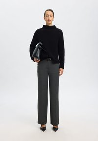 Schwarzer gerippter Pullover mit hohem Kragen, graue maßgeschneiderte Hose und eine schwarze strukturierte Handtasche, dazu spitze schwarze Absatzschuhe.