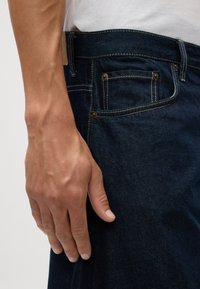 Jeans de mezclilla azul oscuro con costuras amarillas, mostrando un primer plano de la cinturilla, el bolsillo y una mano descansando en el lateral.