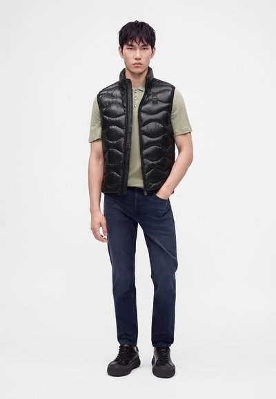 Joven con un chaleco negro acolchado sobre una camisa verde de manga corta, jeans oscuros y zapatillas negras, de pie con una mano en el bolsillo.