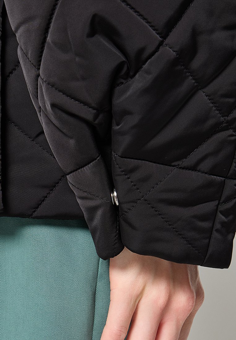 Zwart gewatteerd jack met een gladde textuur, voorzien van een manchetknoop met een zilveren drukknoopdetail, gecombineerd met een lichtgroene stof eronder.