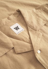Chemise beige à manches courtes avec une texture de lin douce. Présente un col classique, une poche poitrine simple et une étiquette logo blanche. Détail des boutons visible.