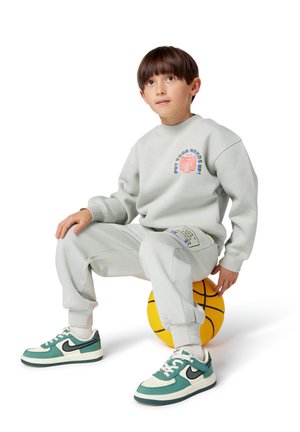 Lichtgrijze sweatshirt met grafisch ontwerp, bijpassende sweatpants en teal-witte sneakers. Zittend op een gele basketbal.