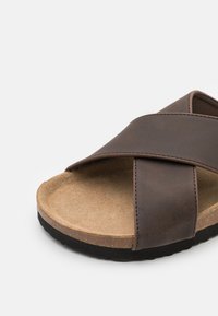 Sandală din piele sintetică maro, cu curele încrucișate, talpă din plută texturată și talpă din cauciuc negru. Designul se concentrează pe minimalism și confort.