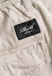 Pantalones de tela beige con una pequeña etiqueta negra que dice "Reell est. 1997". Incluyen costuras prominentes y un detalle de bolsillo con parche.