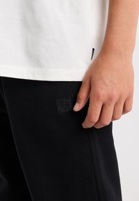White cotton t-shirt and black sweatpants, featuring a fabric tag and a small, textured logo patch on the left leg.