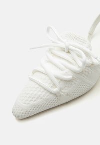 Λευκό μυτερό αθλητικό παπούτσι με υφή από δίχτυ και κορδόνια που διασταυρώνονται δεμένα σε φιόγκο στην κορυφή, εμφανιζόμενο από μια κοντινή γωνιακή λήψη.