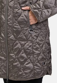 Pikowana, brązowa odzież wierzchnia z teksturowanym wzorem, dwie kieszenie na zamek błyskawiczny oraz zatrzask na dolnym końcu. Charakteryzuje się gładkim, połyskliwym wykończeniem.
