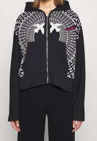 Hoodie zippé noir avec un design graphique en texte blanc et rose accompagné de formes abstraites. Le matériau semble doux et extensible.