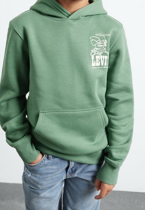 Barn iført grøn Levi's hættetrøje med forlomme og tegneserie-logo, blå jeans, stående med den ene hånd i lommen mod en ensfarvet baggrund.