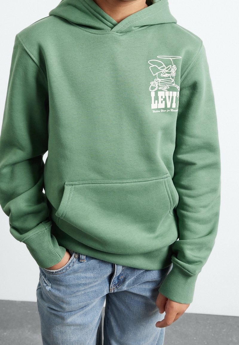 Enfant portant un sweat à capuche vert Levi's avec poche avant et logo cartoon, jean bleu, debout avec une main dans la poche sur fond uni.
