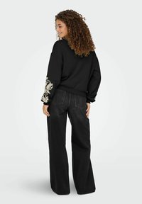 Zwarte crewneck sweatshirt met decoratief beige ontwerp op de linker mouw, gecombineerd met hoog-gerende, wijde zwarte jeans.