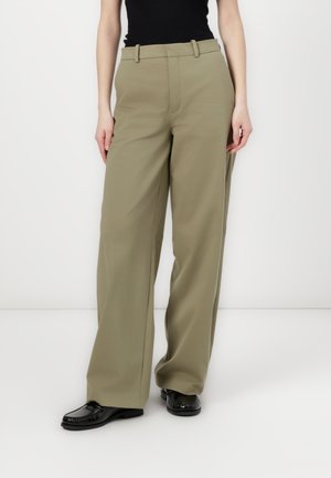 Helle olivfarbene weite Hose aus glattem Stoff, mit Gürtelschlaufen und hoher Taille. Getragen mit schwarzen strukturierten Schuhen.