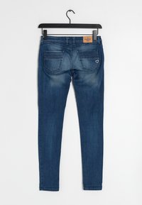 Jeans skinny en denim bleu foncé avec une étiquette en cuir, deux poches arrière, un léger délavage et un détail de couture en forme de cœur sur la poche droite.