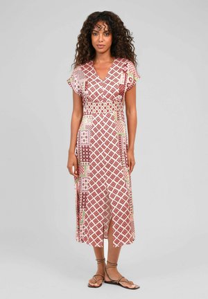 Femme aux cheveux bouclés portant une robe midi rose et rouge à motifs avec manches courtes et fente, debout avec une expression neutre et des sandales.