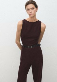 Bordeaux jumpsuit med ärmlös design, texturerat tyg och ett åtsittande svart bälte med en cirkulär spänne vid midjan.