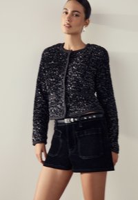 Svart kort jacka med paljetter och långa ärmar, kombinerad med svarta högmidjade jeansshorts med framfickor och ett bälte.