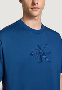 Modrá bavlněná trička s kulatým výstřihem, která má vyšitý logotyp Calvin Klein v tonálním designu. Měkká textura, krátké rukávy a volný střih.