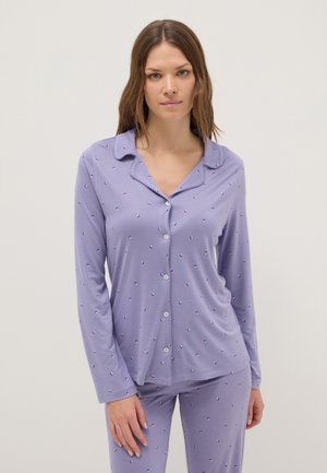 Donna che indossa una camicia da pigiama lavanda con maniche lunghe e bottoni e pantaloni abbinati con piccolo motivo a pois, in piedi davanti a uno sfondo chiaro e semplice.