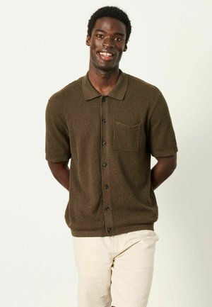 Smiling man wearing a short-sleeve brown button-up shirt with a chest pocket and light beige shorts, standing with hands behind back.