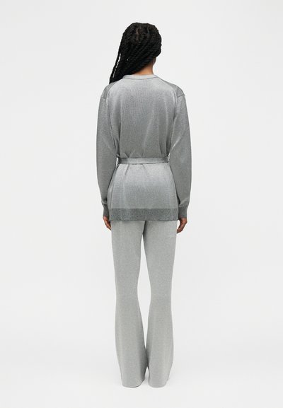 Γυναίκα με μακριά πλεκτή κοτσίδα, φοράει ανοιχτό γκρι ζακέτα με ζώνη και ταιριαστό φαρδύ παντελόνι, στέκεται με την πλάτη προς το μέρος μας σε λευκό φόντο.