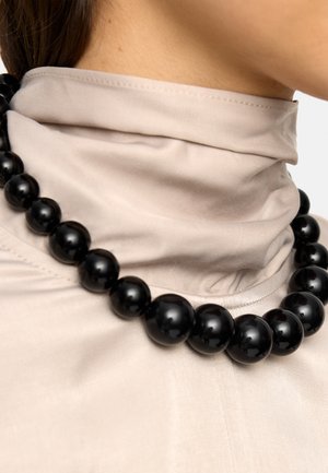 Kobieta w beżowej bluzce z wysokim kołnierzem i masywnym czarnym naszyjniku z koralików spoczywającym na obojczyku.