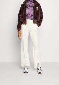 Brązowa kurtka na zamek z futerka syntetycznego, fioletowa bluzka w wzory z d�ługim rękawem, białe szerokie spodnie z rozcięciami oraz sportowe buty z czarnymi akcentami.